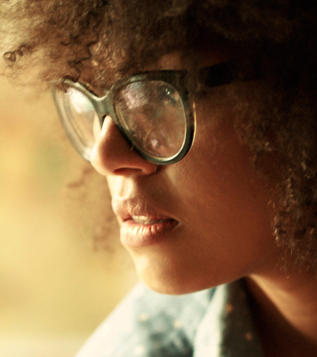 Portrait in warm natural light — close profile of a woman with an afro and oversized round glasses, backlit in golden tones.