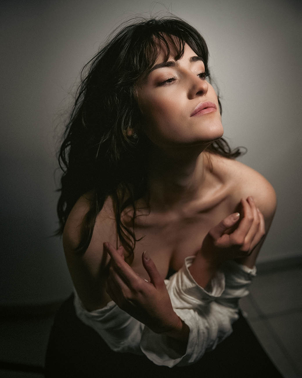 Studio portrait in muted tones — a woman with dark wavy hair tilts her head back, eyes closed, hands drawn to her collarbone.