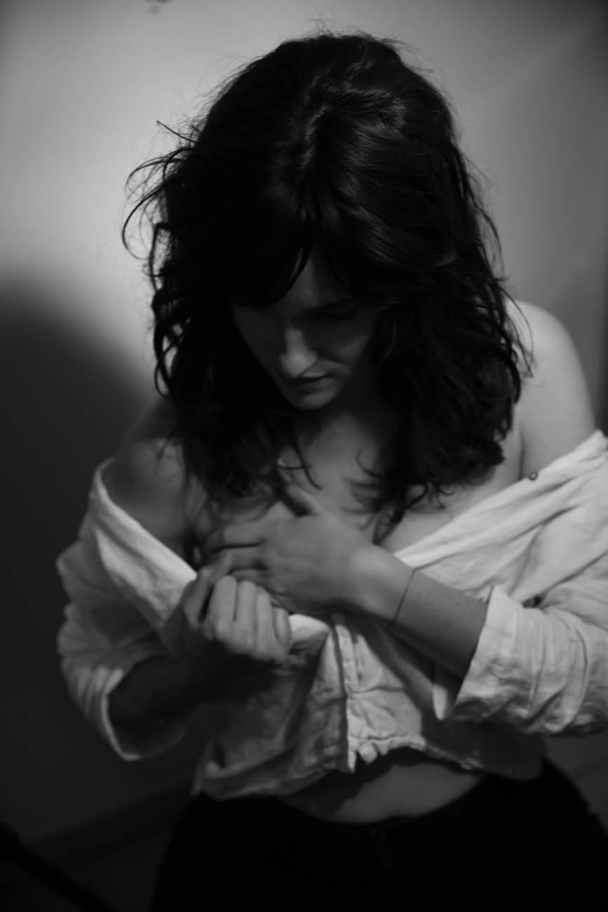 Black and white studio portrait — a woman looks down, dark curls falling forward, an open white shirt slipping from her shoulders.