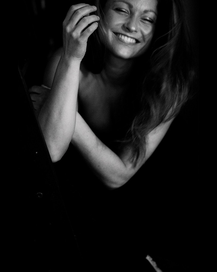 Black and white portrait in deep shadow — a woman laughing with her eyes closed, one hand in her hair, the frame almost dark around her.