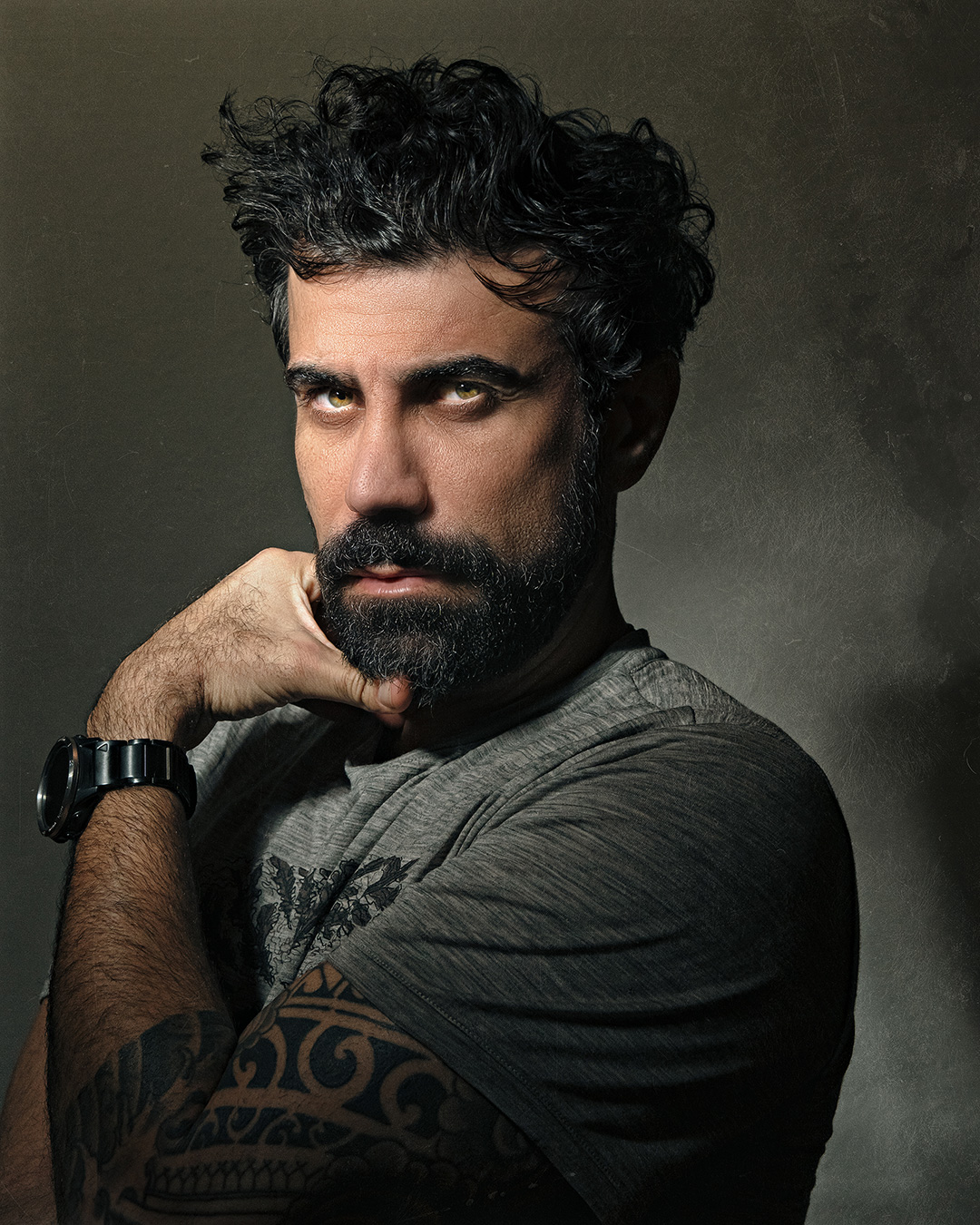 Studio portrait of a bearded man against a grey backdrop — chin resting on his hand, forearm tattoo visible, looking straight into the camera.