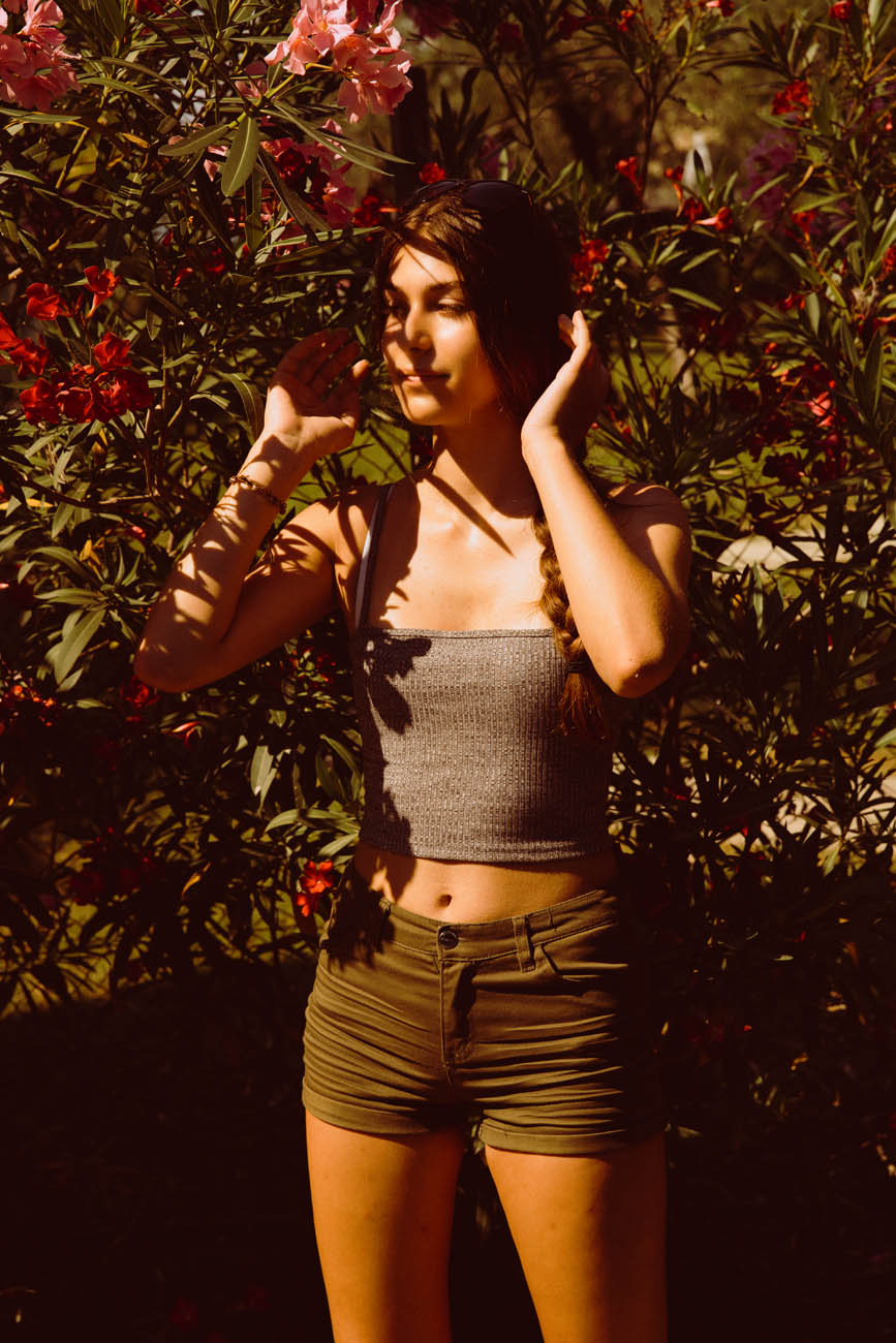 Outdoor portrait in hard afternoon sun — a young woman in front of a flowering oleander, dappled shadow across her face and shoulders.