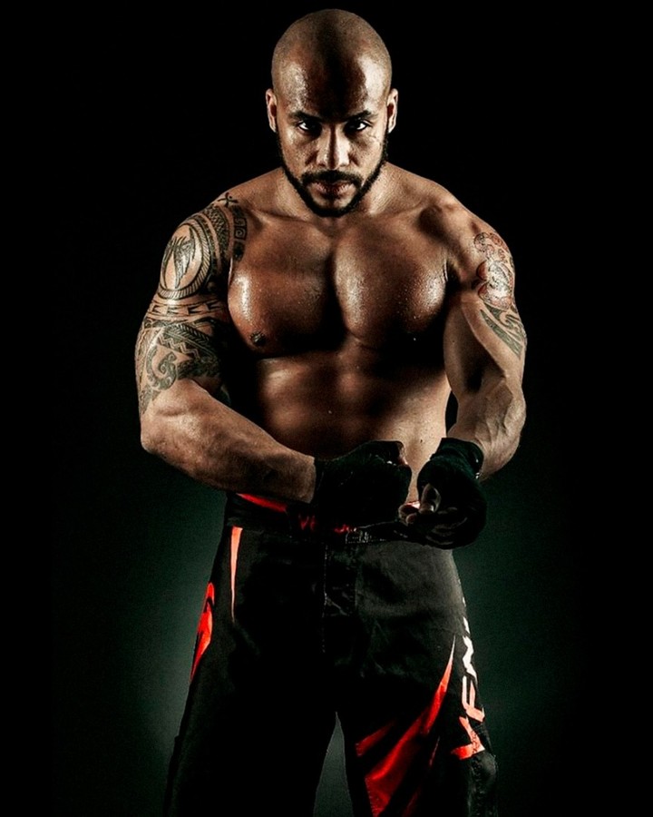 Shirtless, heavily tattooed boxer adjusting his hand wraps under hard rim light against black — sports photograph by Hamudi Leon