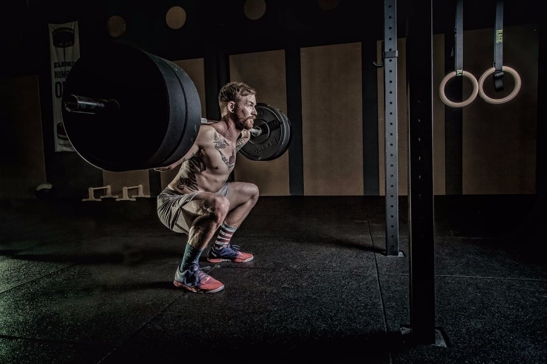 Bearded athlete deep in a back squat under a loaded barbell in a power rack — sports photograph by Hamudi Leon