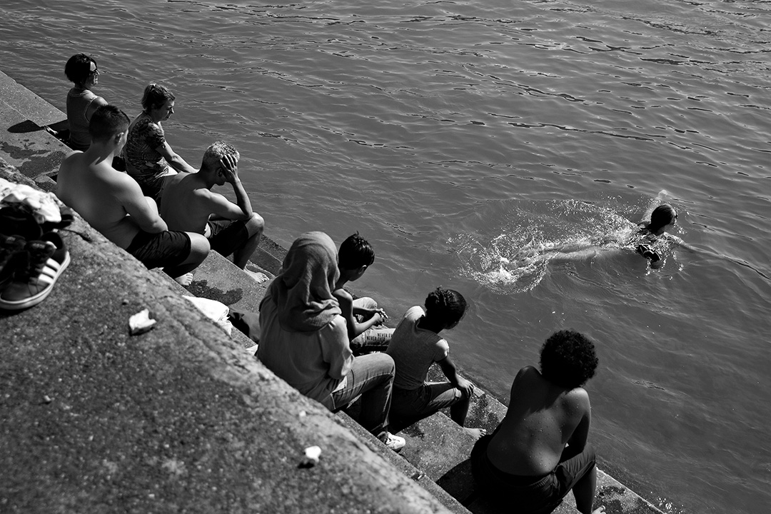 Boys on a stone quay watching one of their own swim in the river below — street photograph by Hamudi Leon