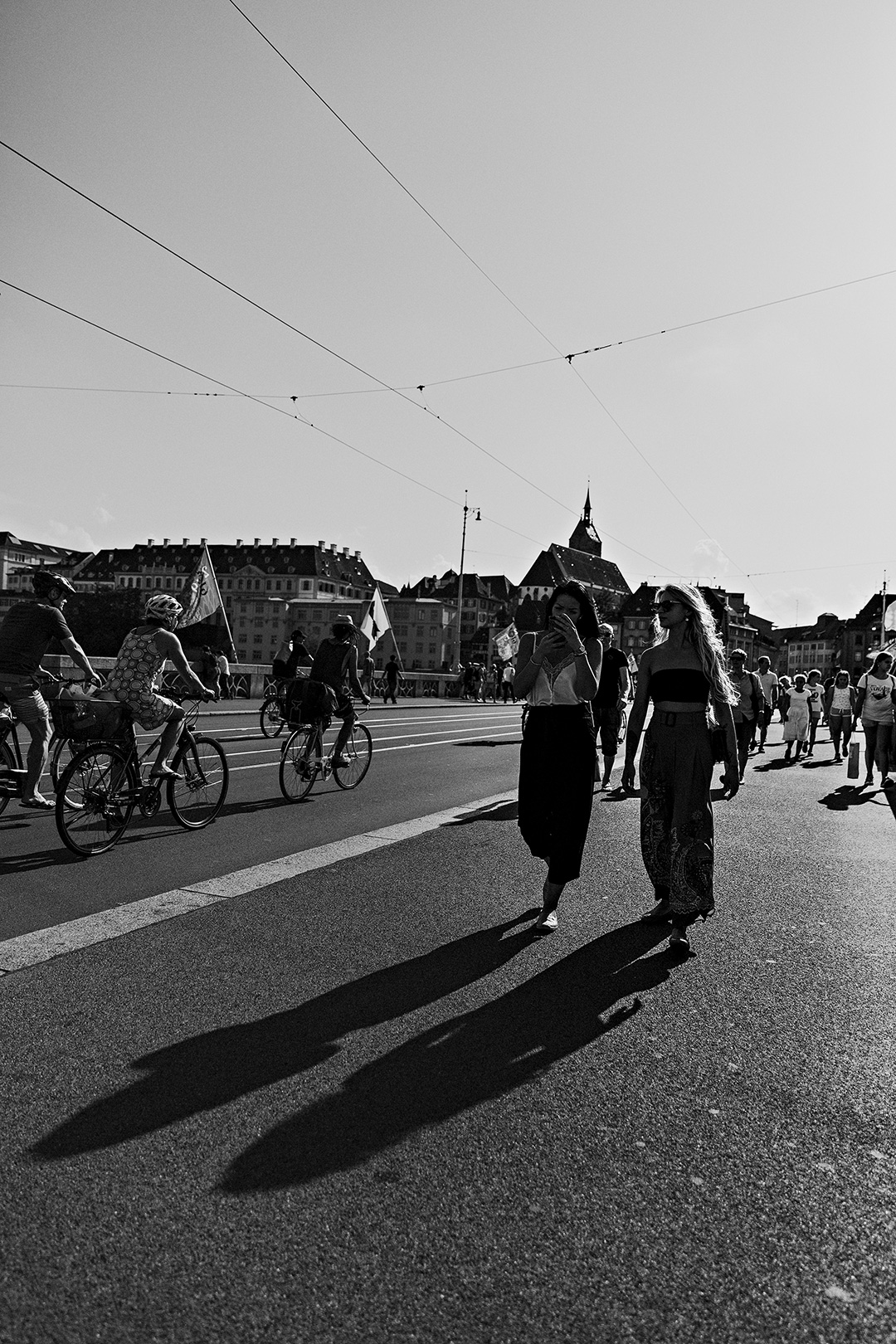 Two people walking a wide sunlit street with long shadows, cyclists and flags behind them — street photograph by Hamudi Leon
