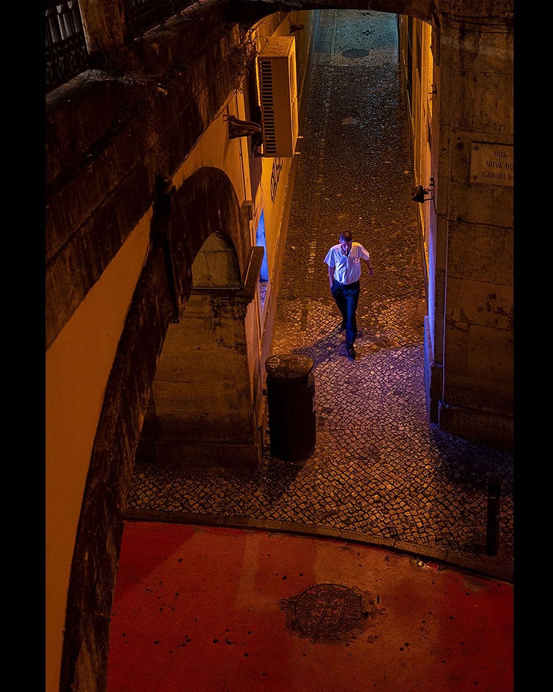 Man walking a narrow alley at night, lit blue and orange from the doorways — street photograph by Hamudi Leon