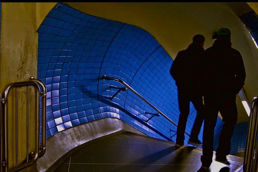 Two figures climbing a stairway lined in blue and yellow tile — street photograph by Hamudi Leon