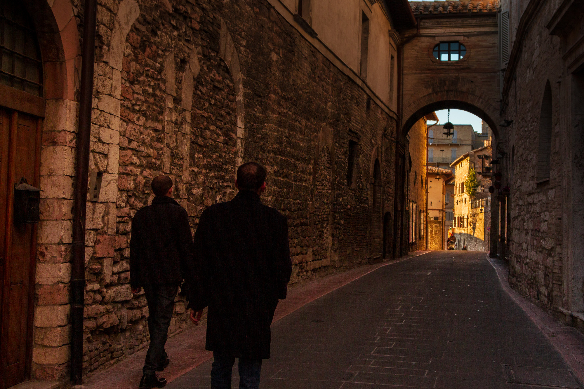 Two men walking a stone street toward an archway — street photograph by Hamudi Leon